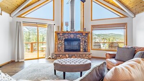 A spacious living room with a stone fireplace, wooden ceiling, and large windows offering mountain views. - Endless View Lodge by Avantstay Incredible Views Close To Bear Mountain Ski Resort w Hot Tub (Big Bear Lake)