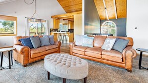 A spacious living area with leather sofas and a tufted ottoman, wooden ceiling beams, and a view of the kitchen and dining area. - Endless View Lodge by Avantstay Incredible Views Close To Bear Mountain Ski Resort w Hot Tub (Big Bear Lake)