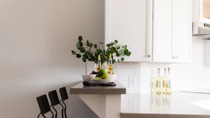 A modern kitchen with white cabinets, a countertop, and a black chair. There is a bowl with fruits and bottles on the counter. - Ivory II By Avantstay Gorgeous Spacious Home w Views Close To Old Town (Scottsdale)