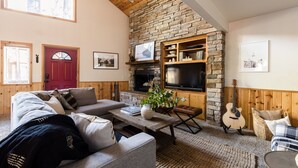 A cozy living room with a stone fireplace, a television mounted on the wall, and a comfortable seating area with a sofa and armchairs. - Jumper By Avantstay Serene Stylish Cabin w Hot Tub (Big Bear Lake)