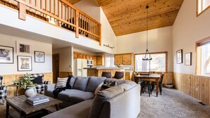 A spacious living area with a large gray sectional sofa, a wooden dining table with chairs, and a kitchen in the background. - Jumper By Avantstay Serene Stylish Cabin w Hot Tub (Big Bear Lake)