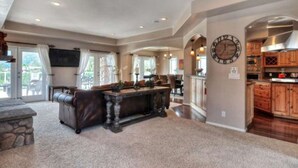 A spacious living room with a fireplace, a large clock on the wall, and a view of the outdoor patio through large windows. - Boulder Bay Lodge by Avantstay Boulder Bay Home w Hot Tub Large Patio Game Room (Big Bear Lake)