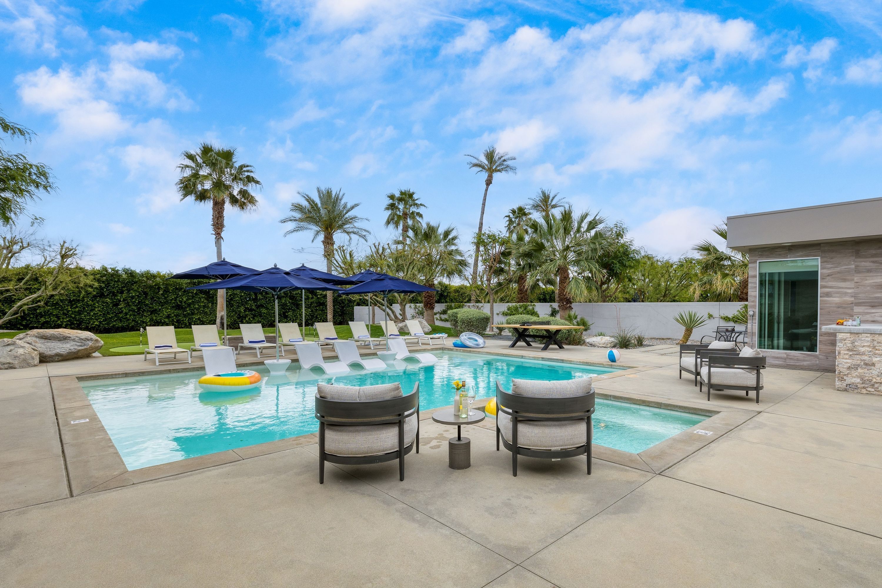 A swimming pool with lounge chairs and umbrellas, surrounded by palm trees and a modern building.