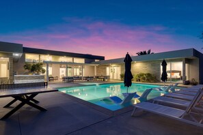 A modern pool area with lounge chairs, a table, and umbrellas, surrounded by a building with large windows and a stone facade. - Polo Villa 10 by Avantstay Backyard Oasis w Putting Green 260320 6 Bedrooms (La Quinta)