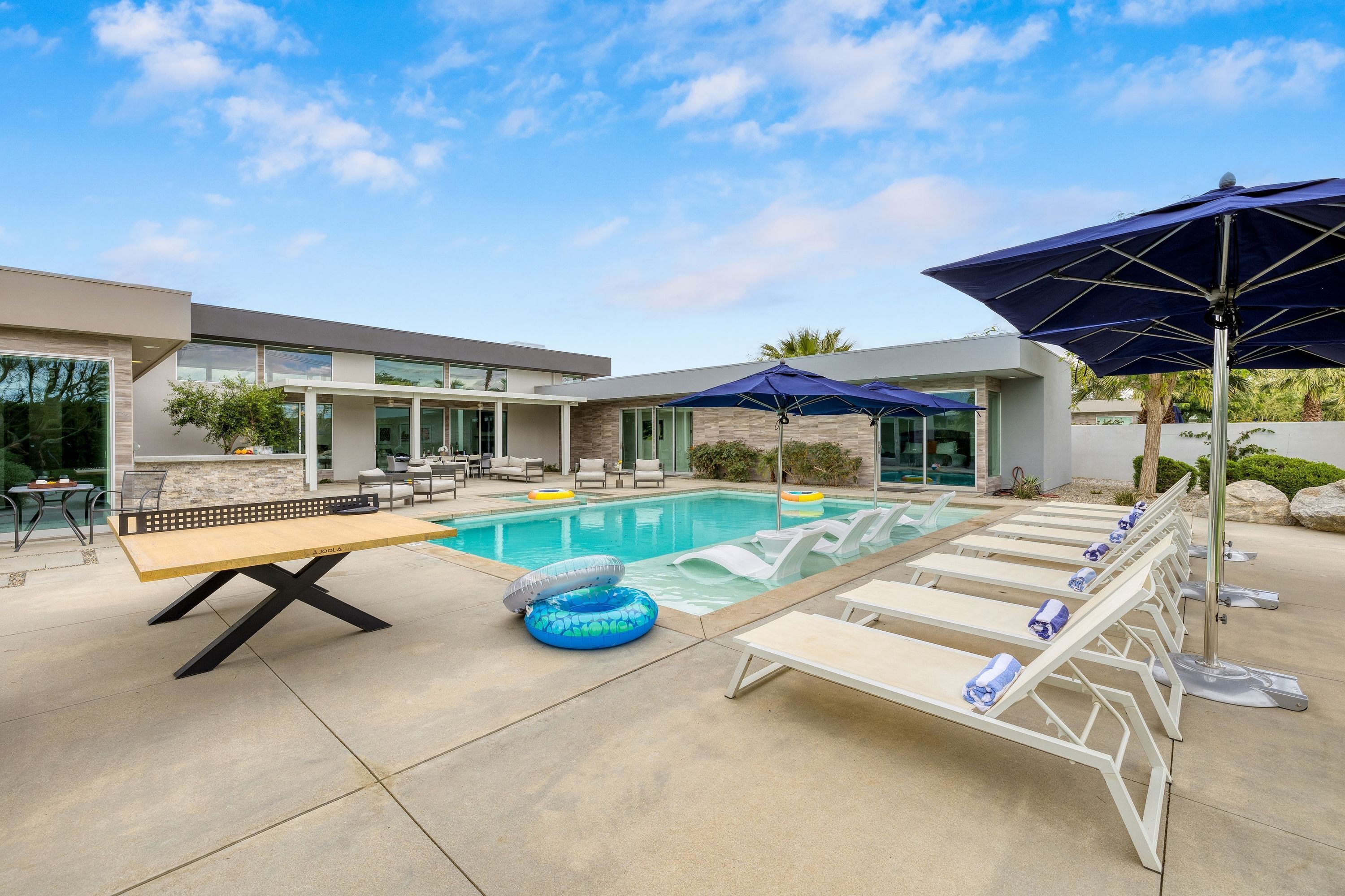 A modern outdoor pool area with lounge chairs, a table, and umbrellas.