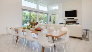 Un comedor moderno con una mesa de madera y sillas blancas, un televisor sobre la chimenea, y grandes ventanas que ofrecen vistas al follaje exterior.