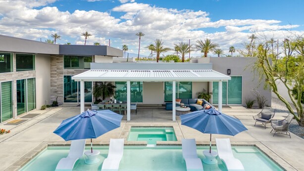 Un área de piscina moderna al aire libre con dos sillones de playa y sombrillas, un espacio de estar techado y una vista de un edificio contemporáneo al fondo.