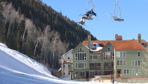 A ski lift with skiers, a green building with multiple windows, and a snowy mountain backdrop. - Etta Place Too 103 by Avantstay Close to Town The Slopes In Complex w Communal Pool Hot Tub (Telluride)