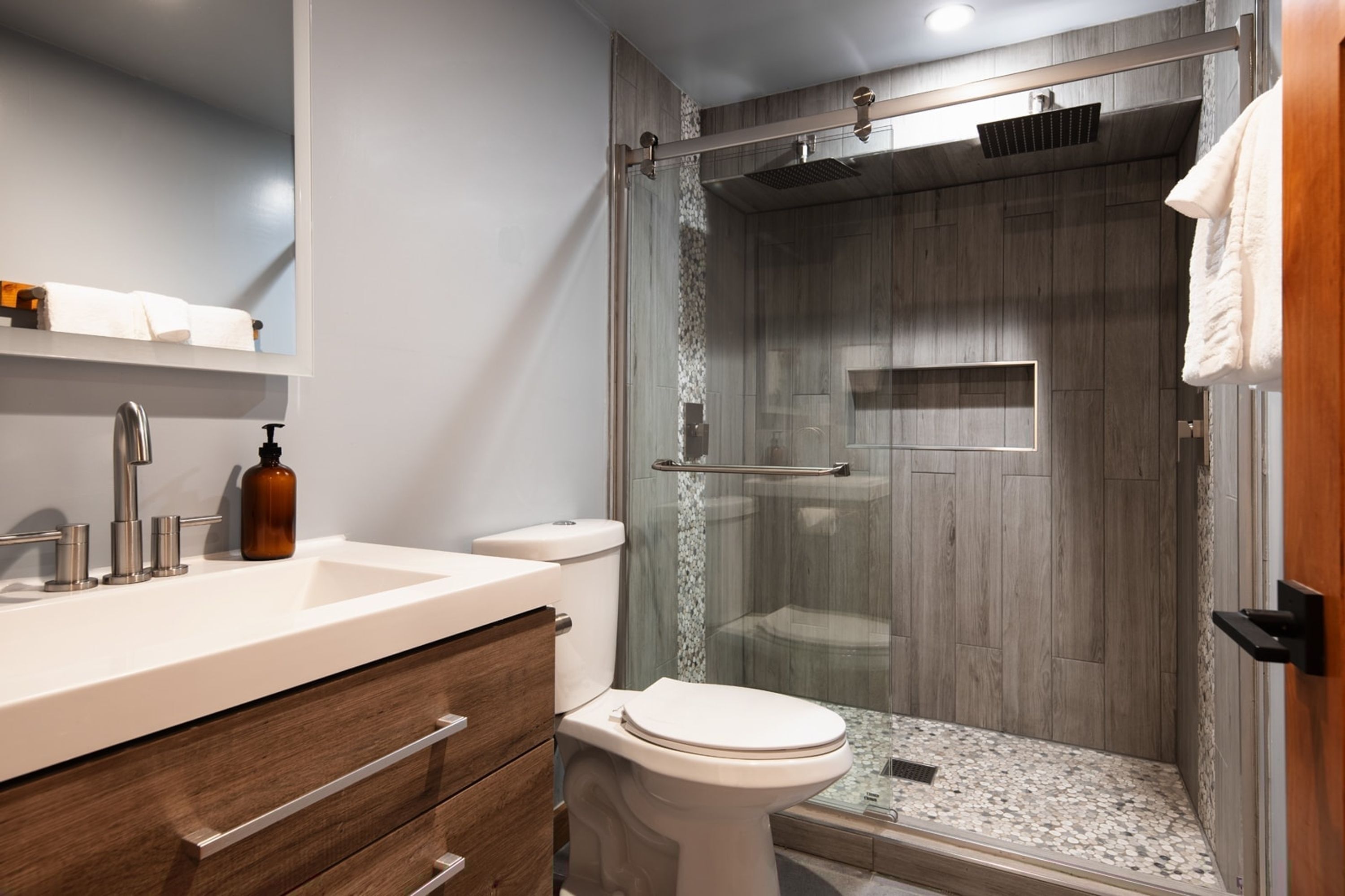 Un bagno moderno con doccia a parete in vetro, lavabo bianco con dispenser di sapone e mobili in legno.