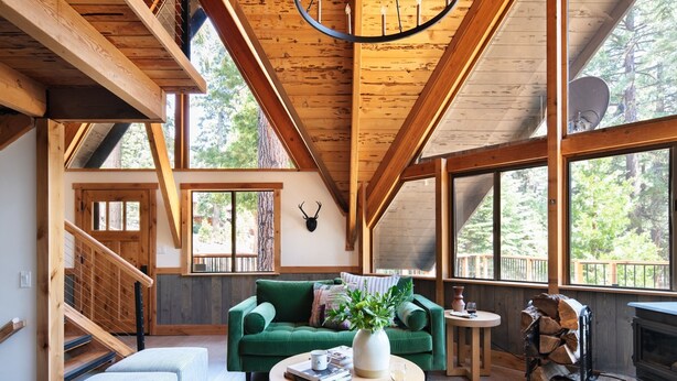 Un vaste espace de vie agrémenté d’un canapé vert, d’une table basse ronde et d’une cheminée. Un escalier mène à un niveau supérieur, et de grandes fenêtres offrent une vue sur la forêt environnante.