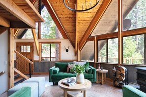 A spacious living area with a green sofa, a round coffee table, and a fireplace. There is a staircase leading to an upper level, and large windows offering views of the surrounding forest. - Timberline by Avantstay Stunning Chalet Cabin w Hot Tub Pool Table Home Theater (Carnelian Bay)