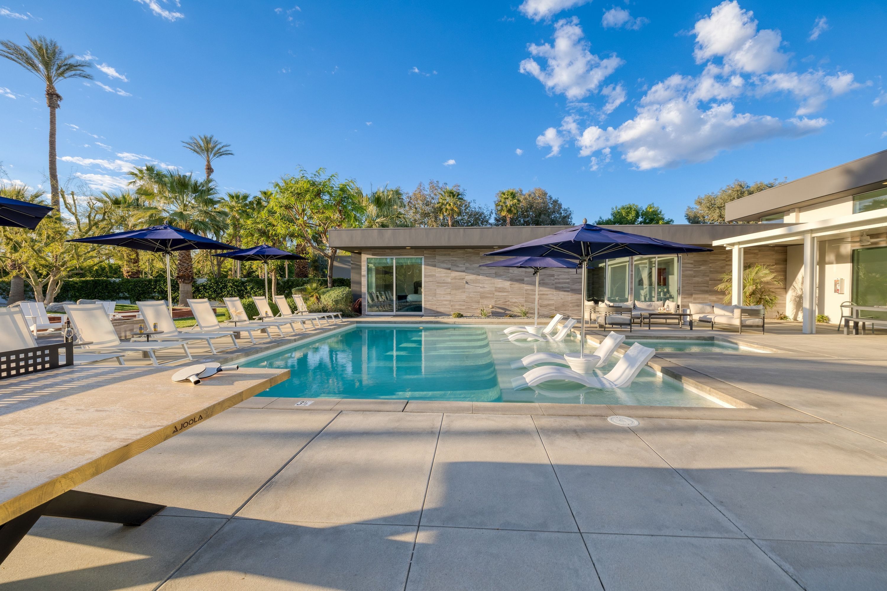 A modern outdoor pool area with lounge chairs and umbrellas, surrounded by palm trees and a clear sky.