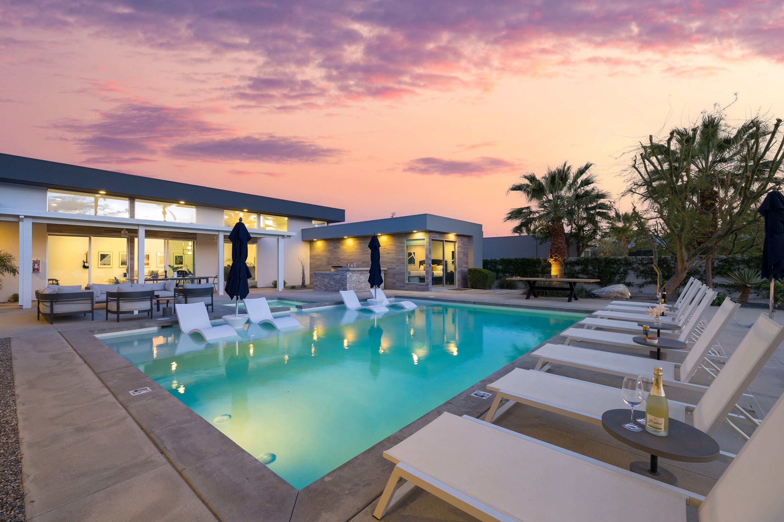 A pool area with lounge chairs and a table with a bottle and glass, surrounded by a modern building with large windows and a sunset sky.