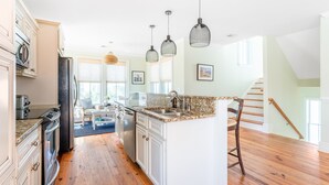 A spacious kitchen with white cabinets, a granite countertop, and a central island. There are hanging pendant lights and a staircase leading to an upper level. - Sand Dune Sea Shell by Avantstay Gorgeous 2 Home Rental w 12 BRs (Isle of Palms)