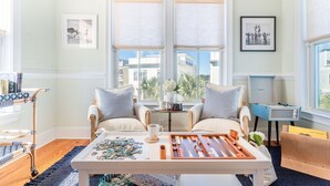 A cozy room with a large window, two chairs, a coffee table with puzzles, and a small table with a teapot and cups. - Sand Dune Sea Shell by Avantstay Gorgeous 2 Home Rental w 12 BRs (Isle of Palms)