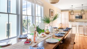 A long dining table set with plates, glasses, and a bottle of wine, surrounded by windows and a kitchen area. - Sand Dune Sea Shell by Avantstay Gorgeous 2 Home Rental w 12 BRs (Isle of Palms)