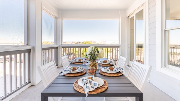 A covered outdoor dining area with a table set for four, surrounded by white railings and large windows. - Sand Dune Sea Shell by Avantstay Gorgeous 2 Home Rental w 12 BRs (Isle of Palms)