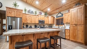 A kitchen with wooden cabinets, a stainless steel refrigerator, a granite countertop, and three bar stools. - Lazy Bear Lodge by Avantstay Pine Tree Paradise w Game Room (Big Bear Lake)