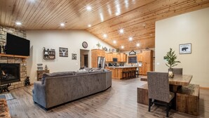 A spacious living room with a gray sectional sofa, wooden flooring, a fireplace, and a kitchen area with wooden cabinets and a dining table. - Lazy Bear Lodge by Avantstay Pine Tree Paradise w Game Room (Big Bear Lake)