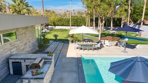 A modern outdoor pool area with a stone barbecue, lounge chairs, and umbrellas. - Polo Villa 6 by Avantstay Features Expansive Putting Green Pool Spa Outdoor Firepit 260324 5 Bedrooms (La Quinta)
