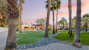 A well-maintained golf course with a putting green, surrounded by palm trees and a modern building in the background. - Polo Villa 6 by Avantstay Features Expansive Putting Green Pool Spa Outdoor Firepit 260324 5 Bedrooms (La Quinta)