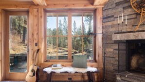 A cozy cabin interior with a stone fireplace, a bench with a fur throw, a guitar, and a large window offering a view of a forest.