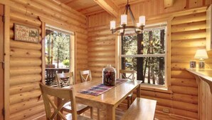 Ein Interieur einer Holzhütte mit Esstisch, Stühlen, einer Chandelüster und großen Fenstern mit Blick auf den Wald.