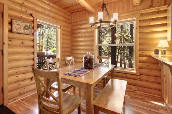 A wooden cabin interior with a dining table, chairs, a chandelier, and large windows offering a view of the forest.