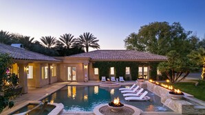 Zona de piscina con sillones de descanso y una chimenea, rodeada de un jardín ajardinado y un edificio con techo de tejas.