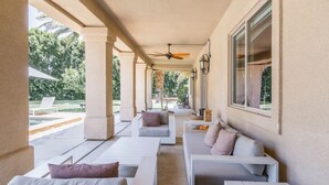 Un espacio de estar al aire libre con techo, con sofás y bancos blancos, un ventilador de techo y vistas a un jardín y una piscina.