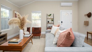 Ein Wohnzimmer mit grauem Sofa, einem Holztisch mit Büchern, einer weißen Vase mit Pampasgras, einem braunen Sessel und einem Bücherregal.