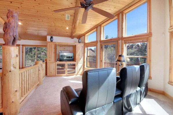 L’intérieur d’un chalet en bois est doté d’un ventilateur de plafond, de grandes fenêtres, d’une cheminée et de fauteuils en cuir.