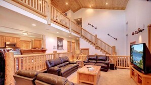 A spacious living room with a large TV, leather sofas, a wooden staircase, and a kitchen in the background. - Starlight Retreat by Avantstay Gorgeous Log Style Home w Incredible Views (Big Bear Lake)