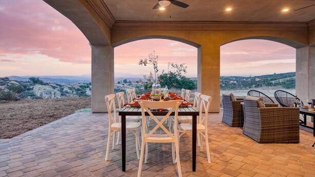 An outdoor dining area with a table set for six, surrounded by white chairs and wicker sofas, overlooking a scenic landscape. - Malbec by Avantstay Incredible Estate w Panoramic Views Bocce Ball Court (Temecula)