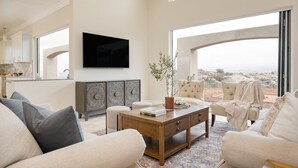 A spacious living room with a large window offering a view of the desert landscape, a flat-screen TV mounted on the wall, and a cozy seating area with a coffee table. - Malbec by Avantstay Incredible Estate w Panoramic Views Bocce Ball Court (Temecula)