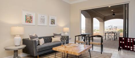 A modern living room with a grey sofa, a wooden coffee table, and two side tables with lamps. There are framed artworks on the wall and a view of an outdoor patio area.