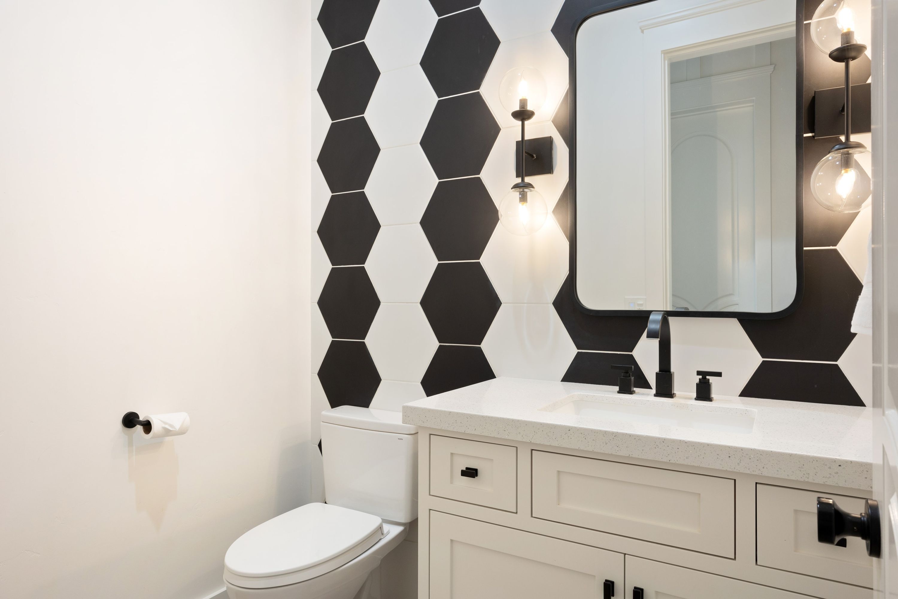 A bathroom with a white toilet, a white sink with a mirror above it, and black and white hexagonal tiles on the wall.