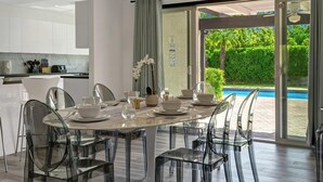 A modern dining area with a glass table and chairs, a view of a swimming pool, and a kitchen in the background. - Catalina by Avantstay Fully Remodeled Palm Springs Haven Pool Permit 3432 (Palm Springs)