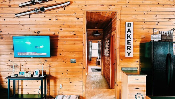 A wooden cabin interior with a television displaying a news channel, a sign reading "BAKERY", and a ski rack on the ceiling. - Cozy 2-bedroom cabin in welcoming Sangerville with WiFi (Sangerville)