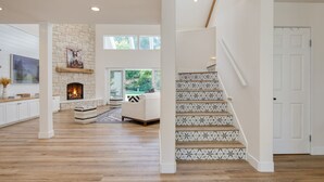 A spacious living area with a fireplace, a staircase with patterned tiles, and a view of greenery outside. - Quarter Horse by Avantstay Countryside Retreat (Temecula)