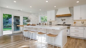 A modern kitchen with a large island, bar stools, white cabinetry, and a view of the outdoors. - Quarter Horse by Avantstay Countryside Retreat (Temecula)