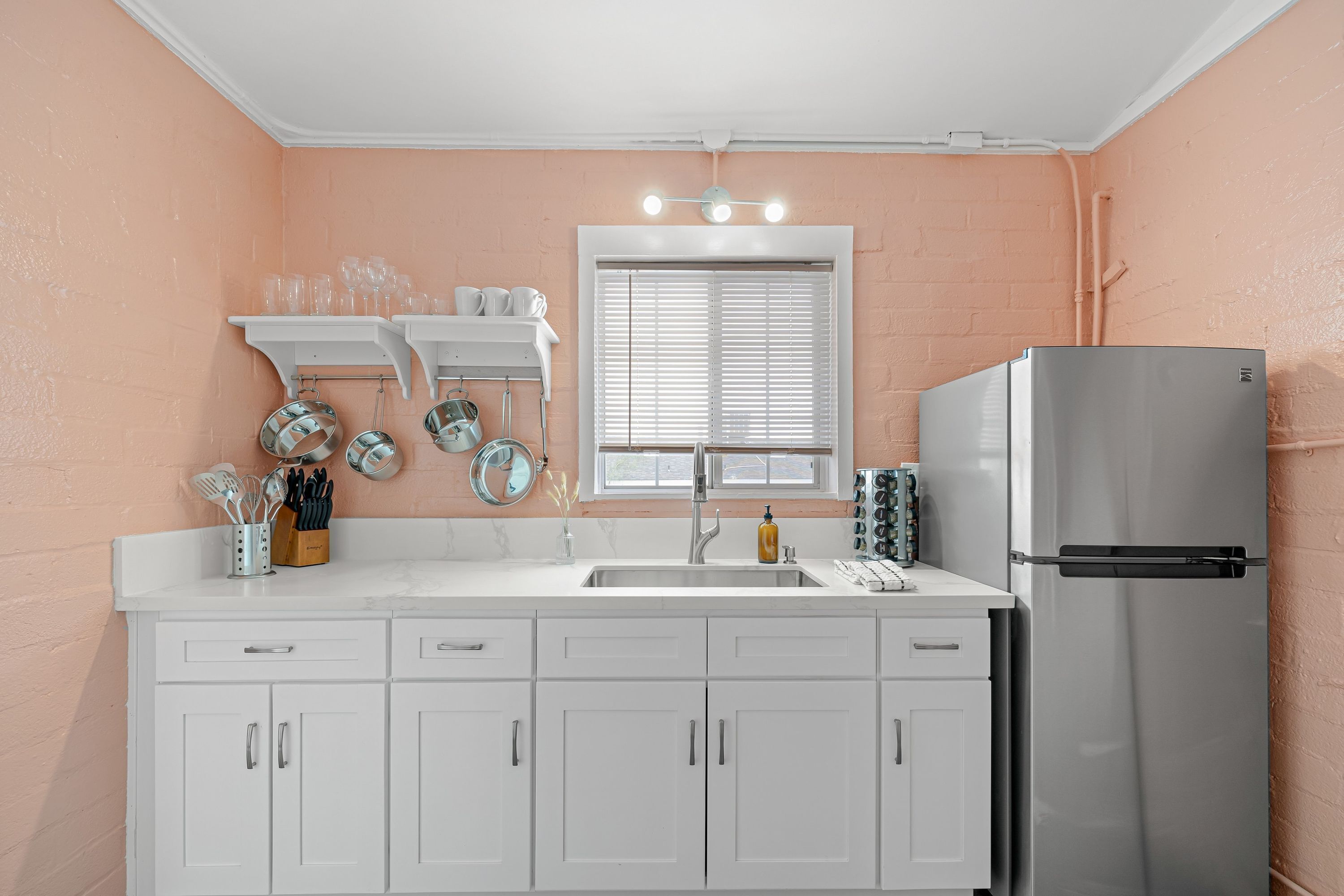 A kitchen with white cabinets, a stainless steel refrigerator, and a window with blinds.