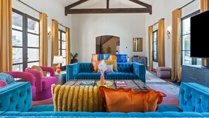 A spacious living room with a large blue sofa, a coffee table, and a television. There are curtains on the windows, and a ceiling fan is visible.