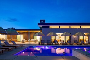Un área de piscina moderna al aire libre con sillones de descanso y sombrillas, un espacio interior bien iluminado con asientos, y una vista despejada de la piscina.