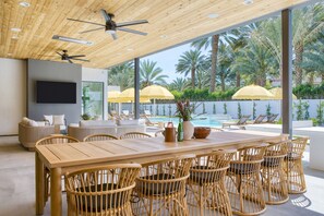 Un amplio espacio exterior para comer con una mesa de madera y sillas de mimbre, con vista a una piscina con sombrillas y palmeras.