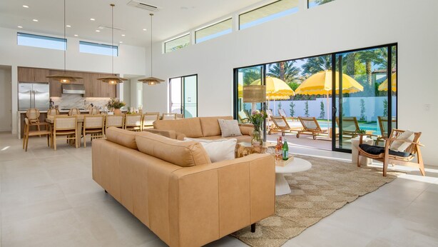 A spacious living area with a large sofa, dining table, and chairs. There is a kitchen in the background, and large windows with a view of palm trees and umbrellas. - The Reserve at Polo Villas 15 by Avantstay (La Quinta)