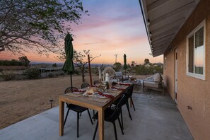 Un espacio de comedor al aire libre con una mesa dispuesta para cuatro personas, rodeada de sillas, con vistas a la puesta de sol y al paisaje desértico.