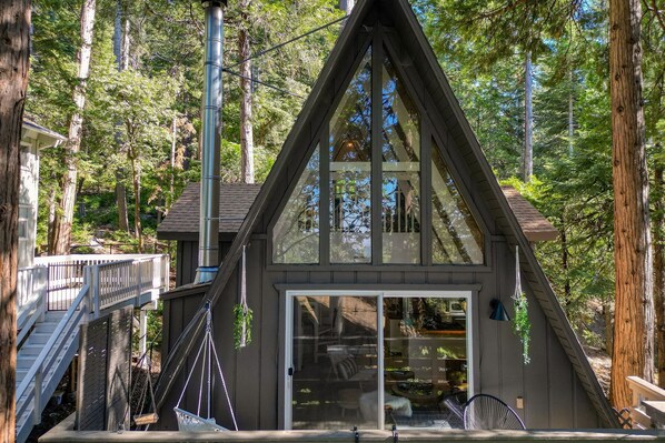 A cabin with a triangular roof, large glass sliding doors, and a deck with a railing. - A-frame Retreat by Avantstay Modern Interiors 2 Decks Views (Skyforest)