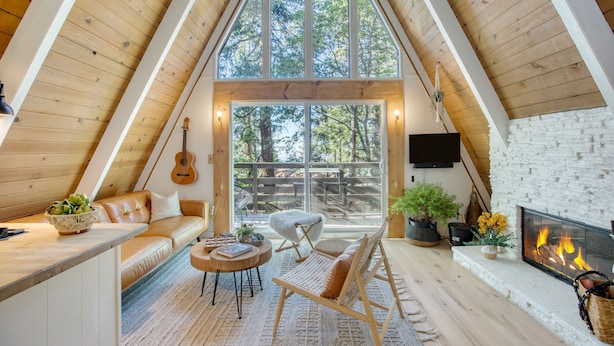 A cozy loft bedroom with a large window, wooden beams, a fireplace, and a guitar on the wall. - A-frame Retreat by Avantstay Modern Interiors 2 Decks Views (Skyforest)