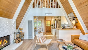 A loft-style living area with a fireplace, wooden floors, a dining area, and a kitchen visible through an open door. - A-frame Retreat by Avantstay Modern Interiors 2 Decks Views (Skyforest)